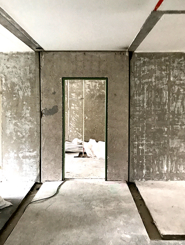 Ein leerer Türrahmen in einem Gebäudeinneren im Rohbauzustand, mit freiliegenden Betonwänden und einem Blick auf Baumaterialien dahinter.