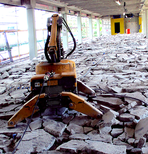 Ein kleiner oranger Abbruchroboter steht inmitten eines großen Haufens von Betonbruchstücken in einem teilweise abgerissenen Gebäude.