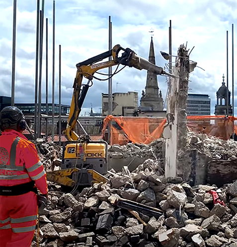Ein Bauarbeiter in roter Arbeitskleidung steht auf einer Baustelle inmitten von Schutt, während ein großer Bagger im Hintergrund ein Gebäude abbricht.