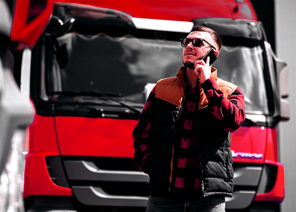 Ein Mann mit Sonnenbrille und einer rot-schwarz karierten Jacke über einer Weste telefoniert vor der roten Front eines Lastwagens. Er blickt dabei leicht nach oben und zur Seite.