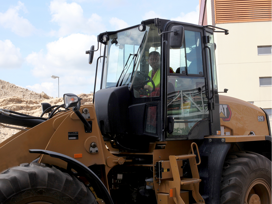 Ein Bauarbeiter in gelber Warnweste sitzt am Steuer eines großen gelben CAT-Radladers auf einer Baustelle unter bewölktem Himmel. Im Hintergrund sind ein Haufen Erdaushub und ein helles Gebäude zu sehen.