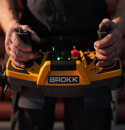 Nahaufnahme der Hände einer Person, die eine gelbe "BROKK"-Fernbedienung mit grünen und roten Lichtern bedient.