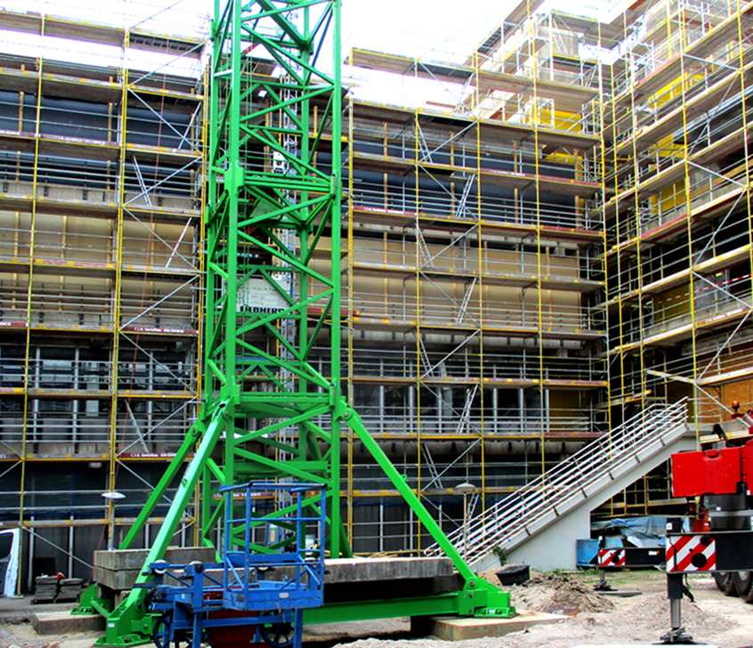Ein hoher grüner Baukran steht auf einer Baustelle vor einem großen Gebäude, das vollständig mit Gerüsten umhüllt ist. Unten links ist eine blaue Hebebühne zu sehen, und rechts führt eine Rampe oder Treppe nach oben.